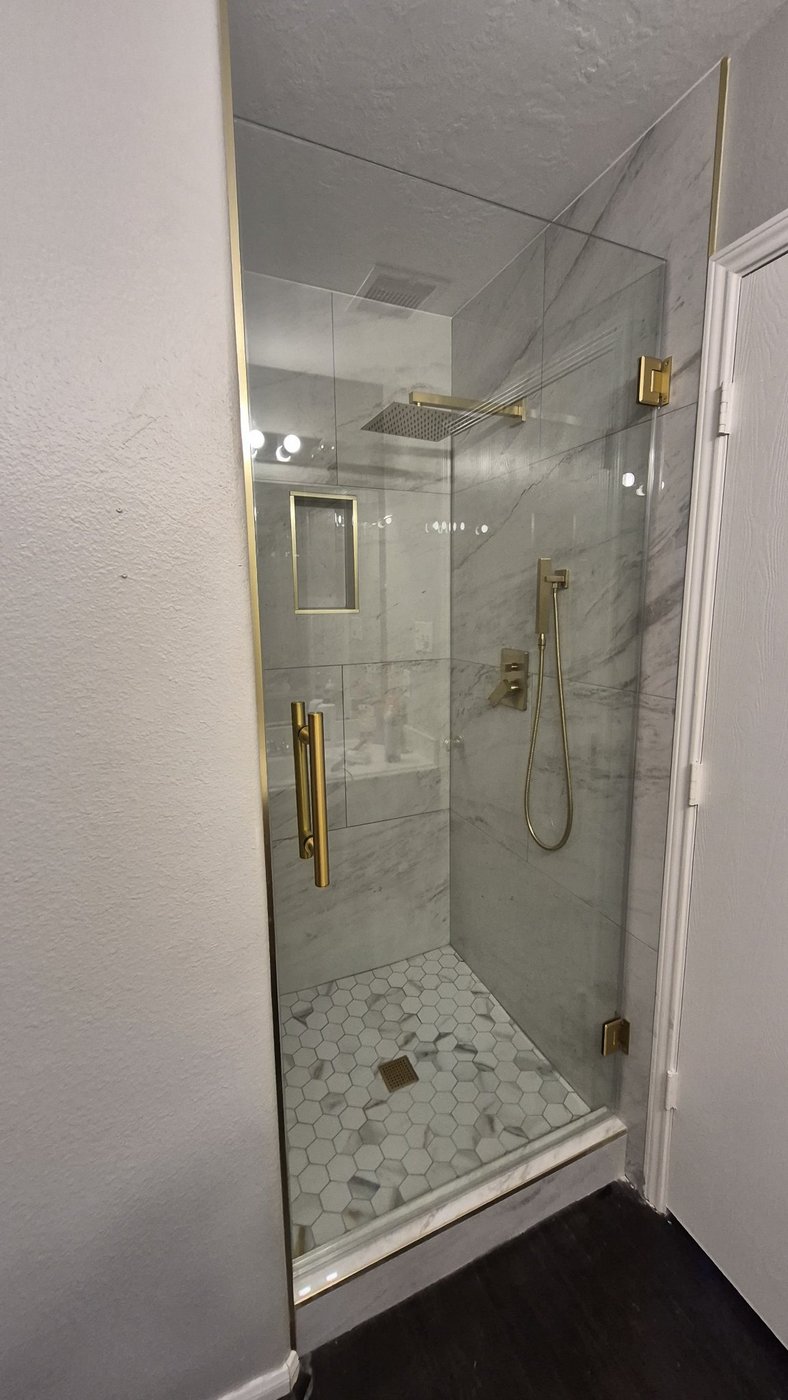 Single door with brush gold hardware installed by Glas Plus in a marble shower with a rain shower head