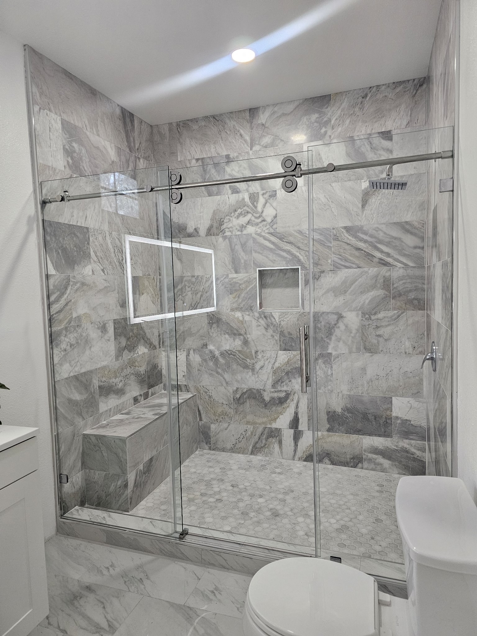 One-door sliding with fixed panel and brush nickel track in a grey marble shower with a bench and niche
